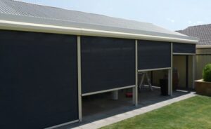 Black mesh outdoor Ziptrak blinds installed on a modern cream pergola overlooking a backyard lawn.