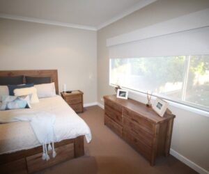 Modern bedroom with a wooden bed frame, matching dresser, and a large window with white roller blinds.
